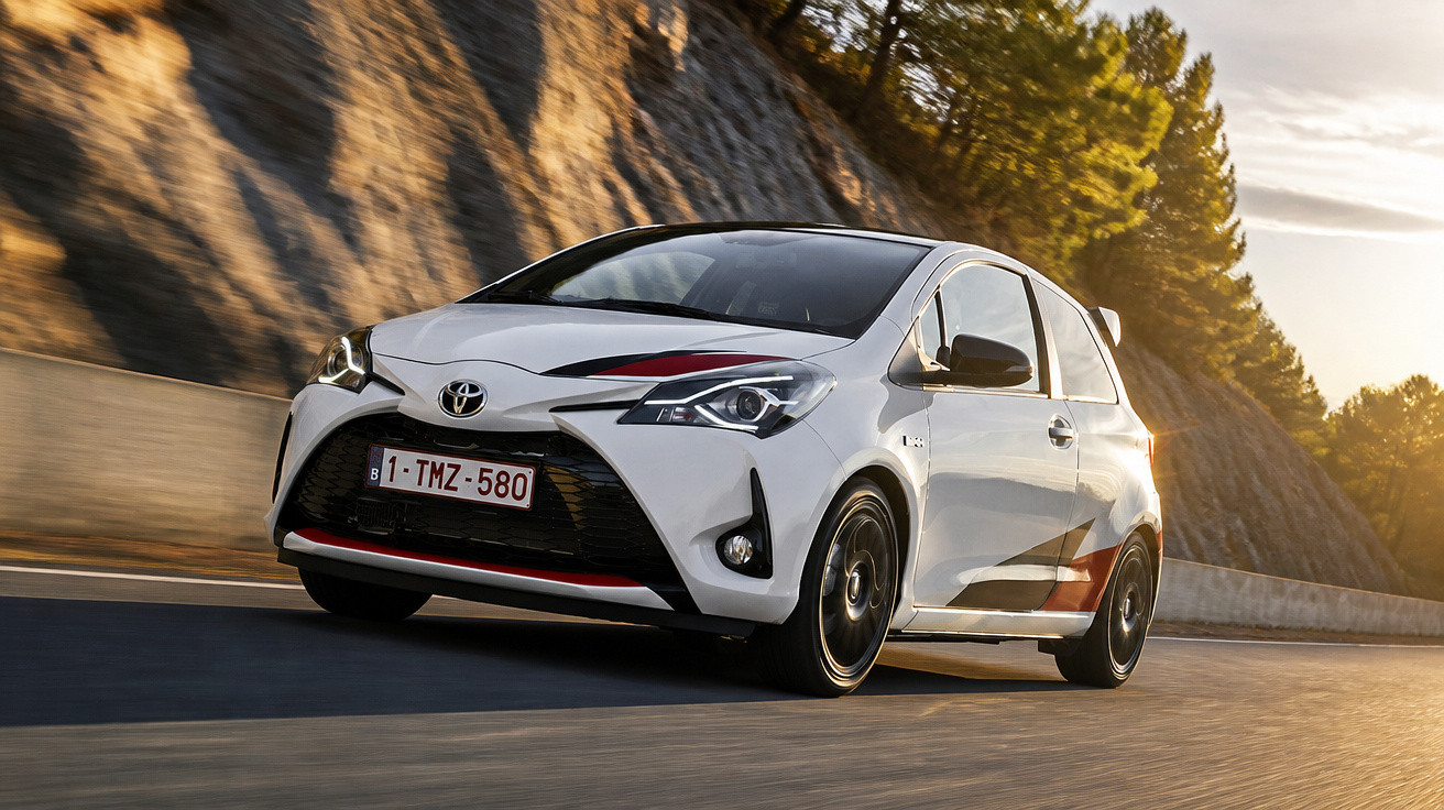 Carro branco Toyota Yaris a circular numa estrada com montanha e árvores ao fundo ao pôr do sol.